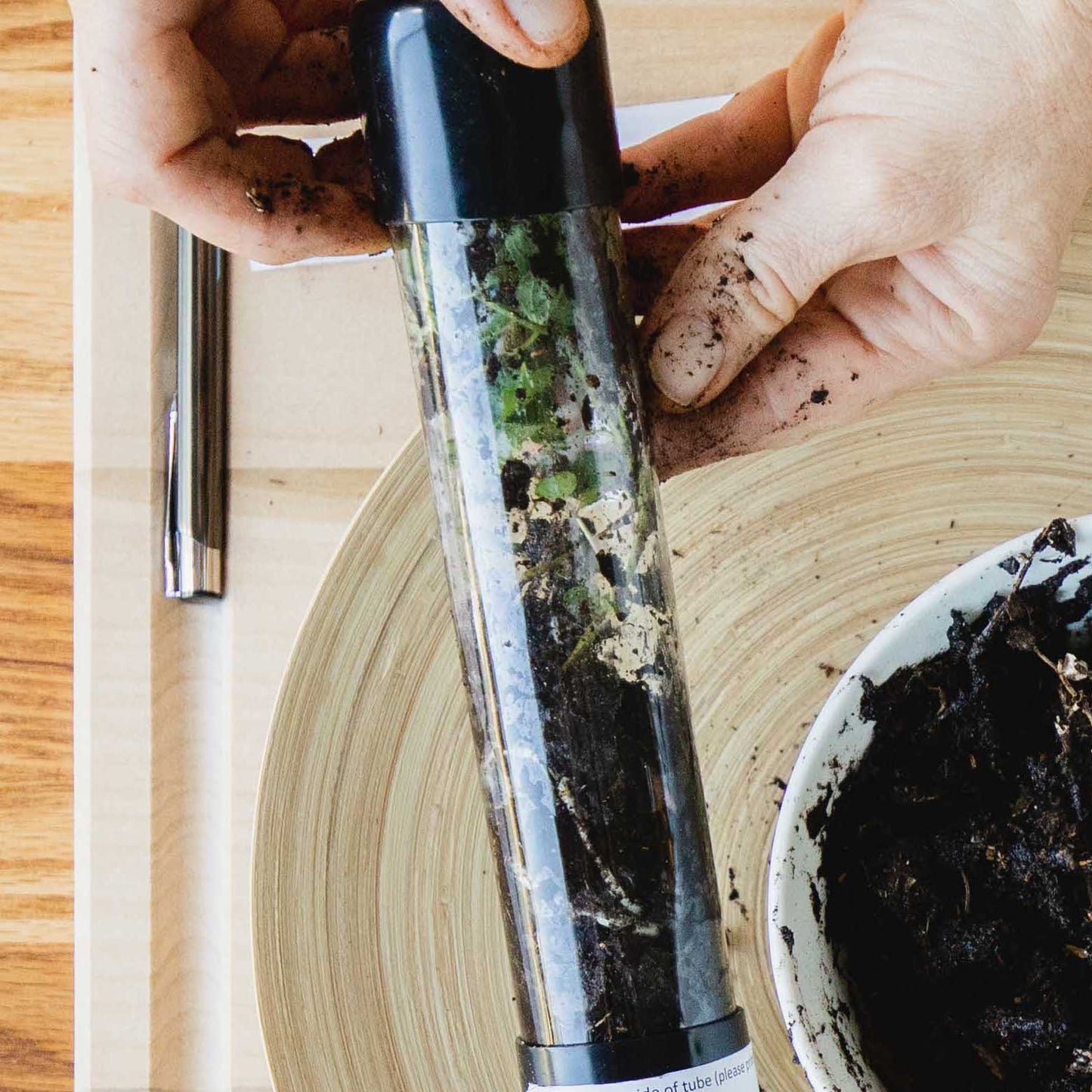 Person holding a plant press tube over a pot with soil, set on a wooden surface.